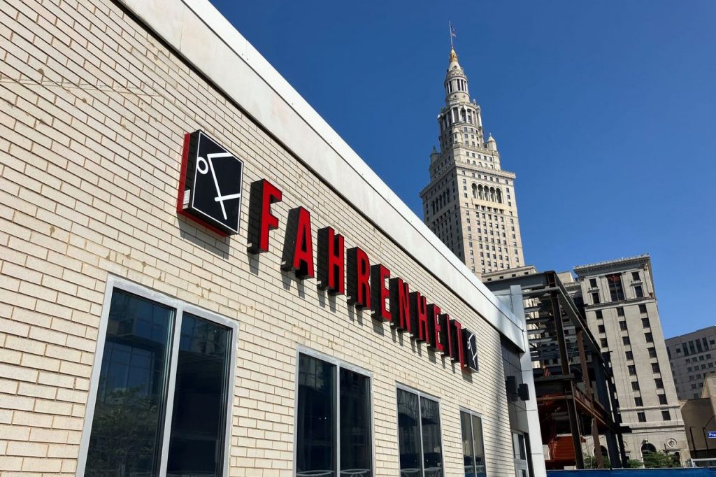The Fahrenheit Cleveland restaurant sign, with its red letters and black logo, is affixed to a cream brick wall on a clear day.