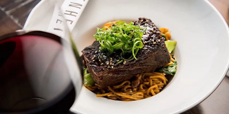A glossy short rib with sesame seeds and scallions rests on a bed of noodles and green vegetables at Fahrenheit Charlotte.