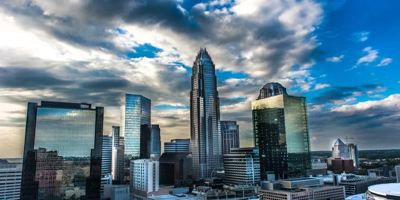 The Charlotte skyline looms under a dramatic cloudy sky, with prominent skyscrapers reflecting the sunlight and clouds.