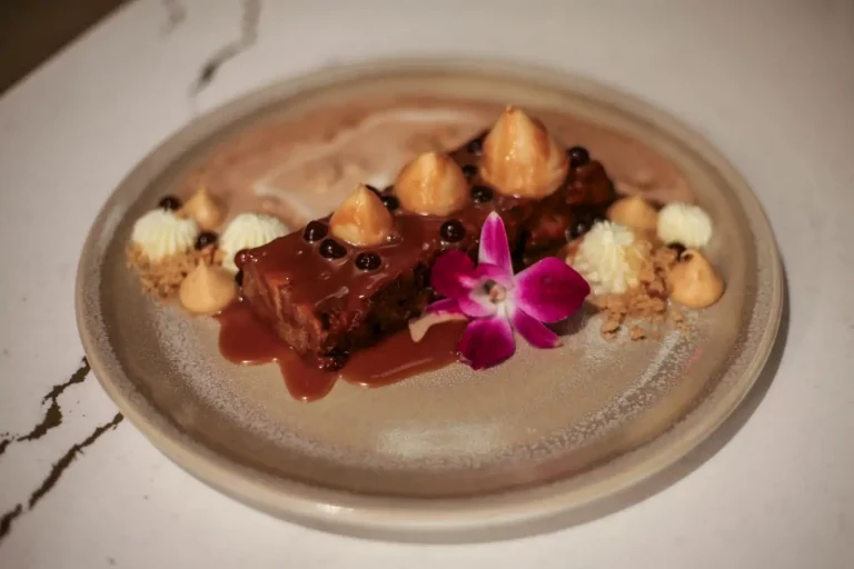 A rectangular piece of bread pudding drizzled with caramel, topped with pearl-like candies, white cream, and an orchid, served on a round ceramic plate at Fahrenheit Cleveland.