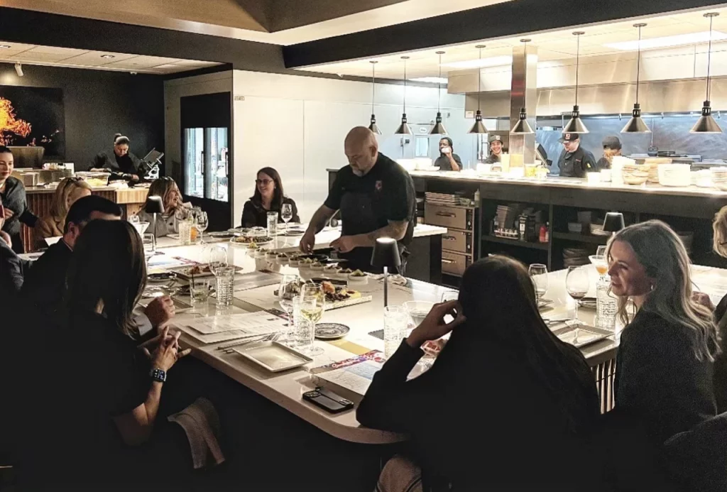 A chef at Fahrenheit Cleveland serves small plates to smiling diners seated at a long, bright counter, with the kitchen visible in the background.
